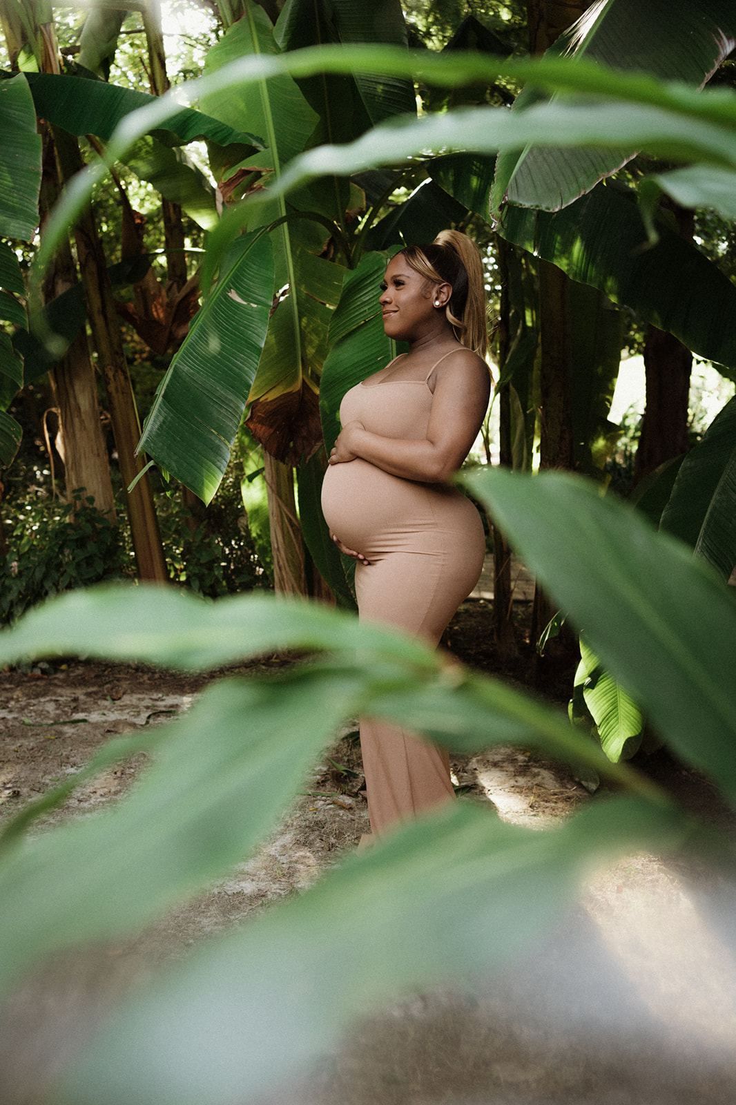 woman stands under large tropical leaves while holding her bump in a brown maternity dress Raleigh OBGYN