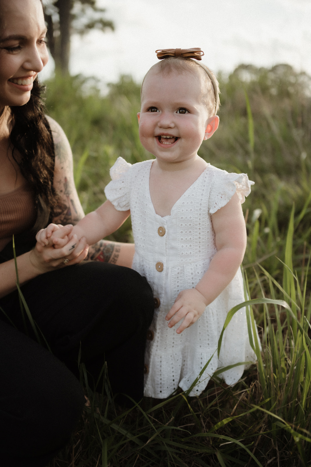 A toddler girl in a white dress plays in a grassy field with her mother