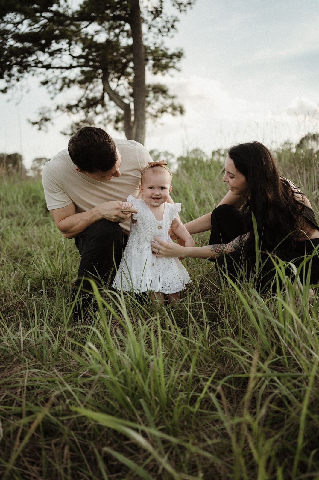 A mother tickles her toddler daughter in a white dress while dad helps her walk through a grassy field Pattywhacks